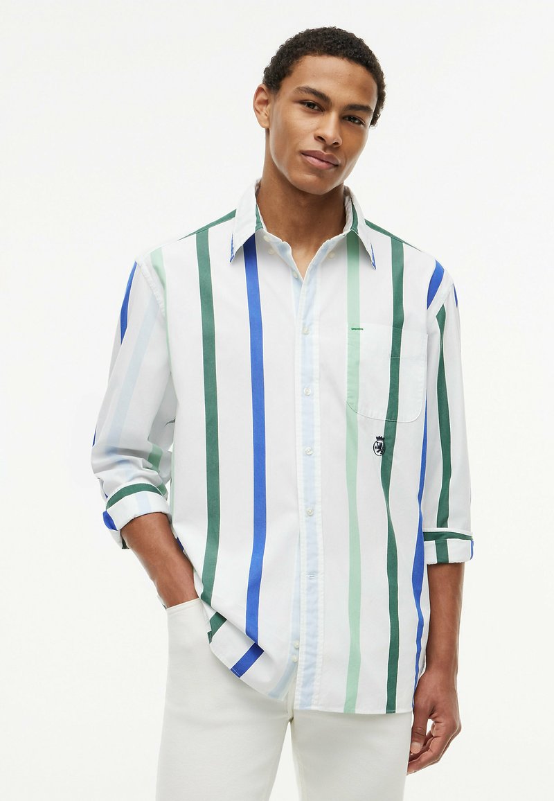 Jeune homme portant une chemise blanche boutonnée avec des rayures verticales bleu, vert et bleu sarcelle, manches retroussées, et pantalon blanc.