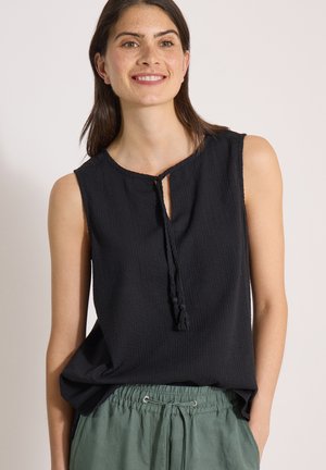 Femme souriante, portant un haut noir sans manches à encolure à nouer et un pantalon vert à cordon, debout devant un fond clair uni.