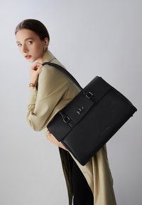 Sort læderhåndtaske med struktureret design, tekstureret finish, dobbelte spænde-detaljer og lang skulderrem, vist mod en lys baggrund.