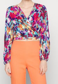 Kobieta nosząca długi rękaw, wielokolorowy kwiecisty top w stylu crop, zestawiony z spodniami w kolorze jednolitym, pomarańczowymi, o wysokim stanie.