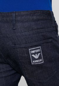 Tmavě modré džíny z denimu s obdélníkovou nášivkou Emporio Armani na pravém zadním kapse, s bílým textem a logem.