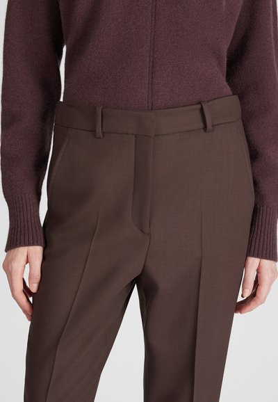 Donna che indossa pantaloni marroni a vita alta su misura e un maglione a coste abbinato, con le mani rilassate lungo i fianchi su uno sfondo semplice.