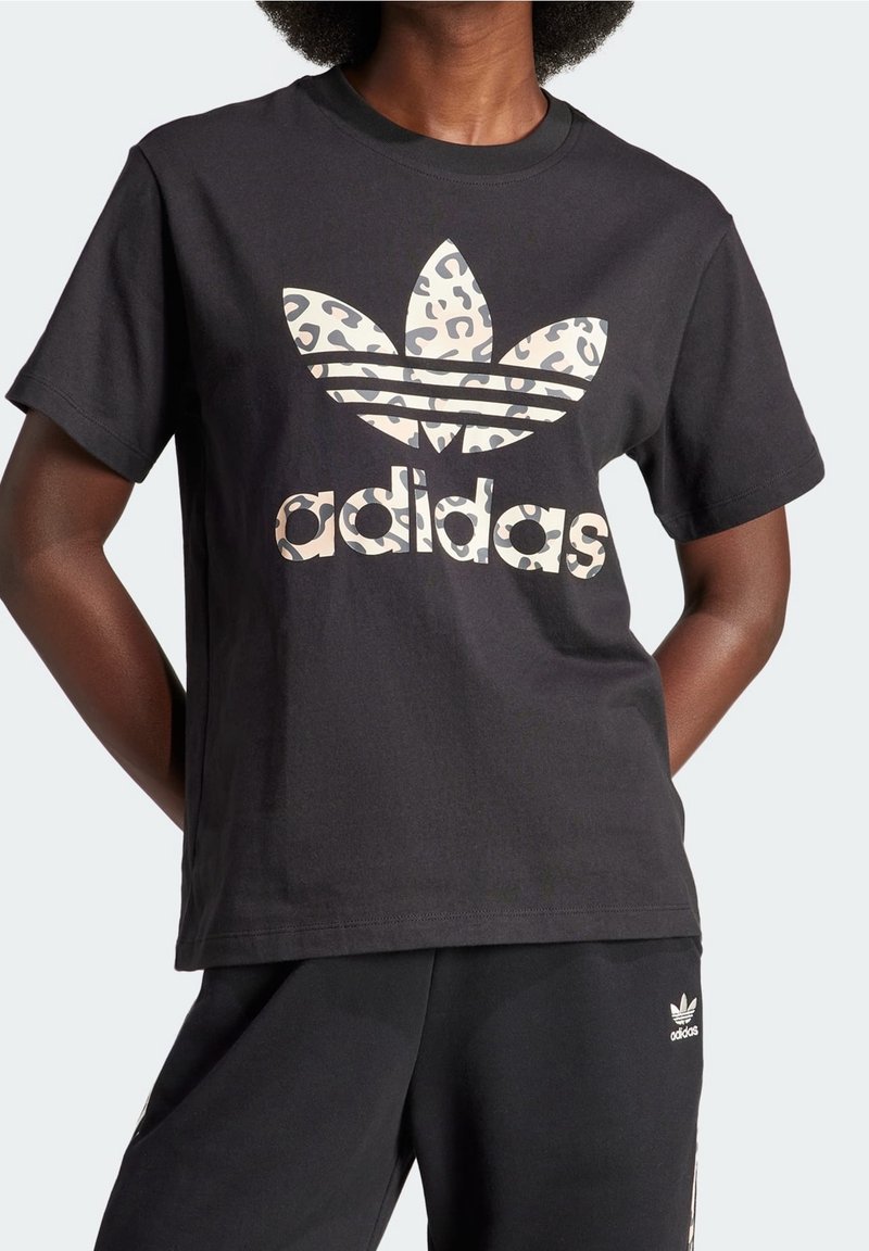Person som bär en svart Adidas t-shirt med leopardmönster och text, tillsammans med svarta Adidas sweatpants.