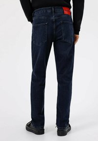 Vaqueros de mezclilla azul oscuro con corte de pierna recta, que cuentan con dos bolsillos traseros y un parche rojo con el logo HUGO en la cintura.