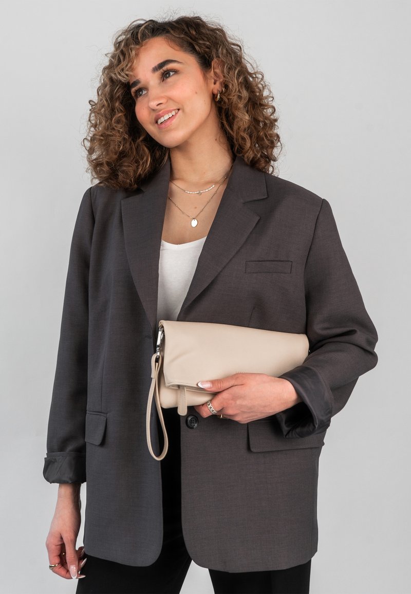 Szürke blézer laza szabással, egyetlen mellzsebbel és strukturált vállakkal. Kézben tart egy bézs bőr táska, amelynek csuklószíja van.