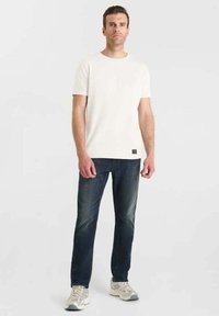Uomo in piedi con una semplice maglietta bianca a maniche corte, jeans blu scuro e sneakers grigio chiaro su uno sfondo bianco.