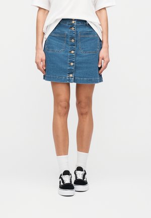 Kvinde iført blå denimknap-front nederdel med forlommer, hvide sokker, sort-hvide sneakers og hvid løs t-shirt.