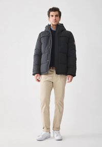 Black puffer jacket with a hood, featuring outer snap closures, side pockets, and a smooth texture, worn over a dark sweater and light beige pants.