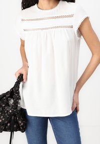 Blusa bianca con maniche corte a scallop, con inserti in pizzo orizzontali e una parte frontale plissettata. Abbinata a jeans blu e una borsa nera floreale.
