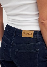 Jeans de mezclilla azul oscuro con un acabado texturizado, que cuentan con una etiqueta de cuero marrón con la inscripción "NA-KD" en la cinturilla y los bolsillos traseros.