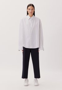 Camicia a righe con una vestibilità ampia, caratterizzata da una base bianca e sottili righe verticali blu navy. Abbinata a pantaloni scuri e sneakers bianche.