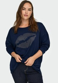 Granatowa koszula z długim rękawem o luźnym kroju z nadrukiem ust ozdobionym cyrkoniami. Gładka faktura, swobodny styl, zestawiona z ciemnymi dżinsami.