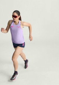 Ljubičasti bezrukavni sportski gornji deo s ružičastim logom, uparen s tamnoplavim šortsem s ružičastim akcentima. Nošeno uz sunčane naočale i sive tenisice za trčanje.