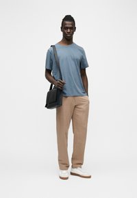 Νέος άνδρας που φοράει μπλε μπλουζάκι, μπεζ παντελόνι, λευκά αθλητικά παπούτσια, κρατώντας μαύρη τσάντα ώμου, στέκεται μπροστά σε απλό φωτεινό φόντο.