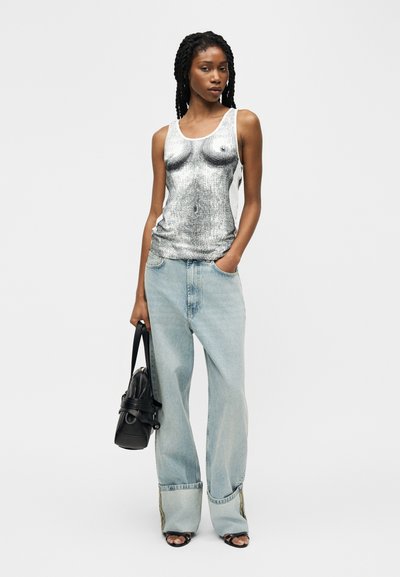 Νεαρή γυναίκα με πλεξούδες, που φοράει αμάνικη μπλούζα με μονόχρωμο τύπωμα στο κορμό, φαρδιές ανοιχτόχρωμες μπλε τζιν, μαύρα σανδάλια και κρατάει μαύρη τσάντα χειρός.