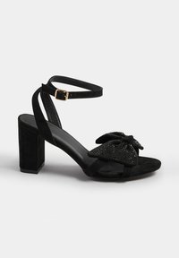 Black suede heeled sandals with a large sparkly bow detail on the toe, featuring a buckle ankle strap and a block heel for stability.