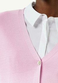 Jasnoróżowy sweter na drutach z dekoltem w serek, zapinany na białe guziki. Noszony na białej koszuli z kołnierzykiem.