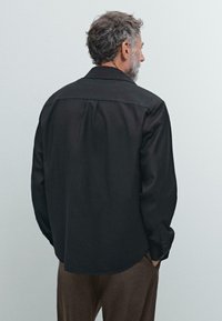 Camicia nera a maniche lunghe con colletto, taglio dritto e polsini con bottone. Texture morbida e design minimale, caratterizzata da una discreta piega sul retro.