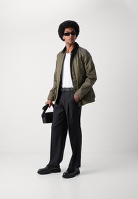 Chaqueta verde oliva, camiseta blanca, pantalones negros y zapatos. Sosteniendo un bolso negro; usando gafas de sol oscuras. Texturas suaves, corte moderno.