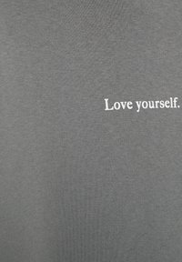 Szary materiał w tle z białym napisem "Kochaj siebie." lekko przesuniętym w prawo.