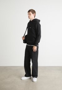 Zwarte hoodie met een kangoeroezak, gecombineerd met losse zwarte broeken en witte sneakers. Model houdt een kleine zwarte tas vast.