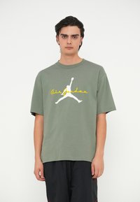 Ελαιόχρωμο βαμβακερό μπλουζάκι με στρογγυλή λαιμόκοψη, διαθέτει λευκό λογότυπο Jumpman και κίτρινο κείμενο "Air Jordan" στο μπροστινό μέρος. Κοντομάνικο.