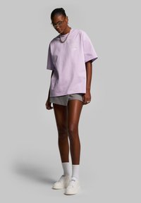 Fialové oversize tričko s krátkými rukávy a detailem loga, zkombinované se šedými šortkami a bílými teniskami. Modelka stojí s uvolněnýma rukama.