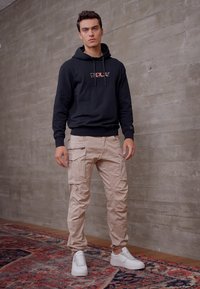 Sort hoodie med logo, beige cargo bukser med flere lommer, og hvide sneakers, sat foran en struktureret grå væg.