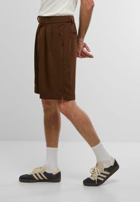 Braune, maßgeschneiderte Shorts mit Falten, seitlichen Taschen und gespaltenem Saum, kombiniert mit marineblauen Sneakers und weißen Socken.