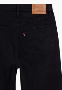 Jean en denim noir avec une pièce en cuir, des coutures de poches arrière courbées et une petite étiquette rouge sur la poche arrière.