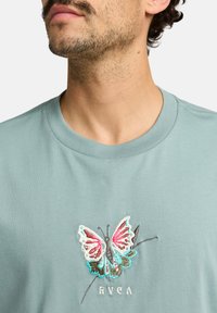 Lyseblå bomulds t-shirt med en brodersyet sommerfugl i pink, hvid og blå, med mærkenavnet "RVCA" under designet.