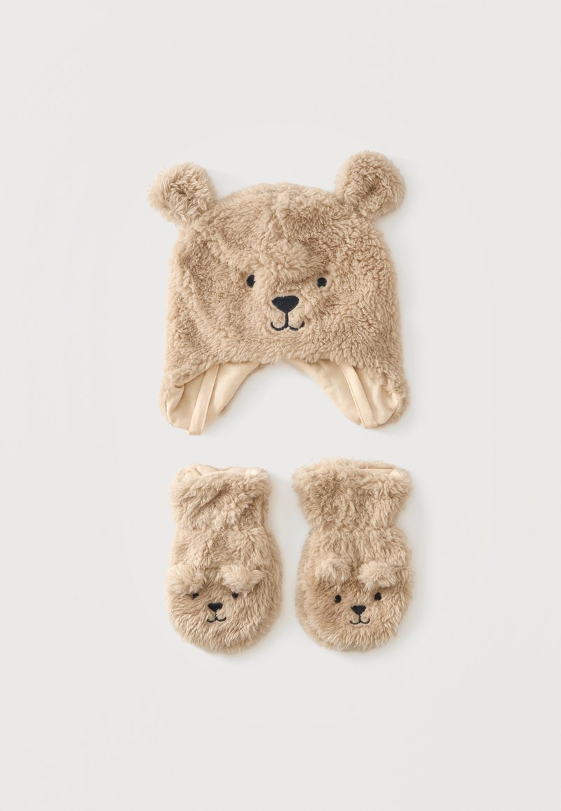 Chapeau beige duveteux avec visage et oreilles d’ours, assorti à des moufles présentant de petits visages d’ours sur un fond blanc uni.