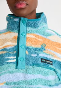 Pullover de fleece com um colarinho alto, apresentando um padrão multicolorido em azul, laranja e creme. Bolso frontal com zíper e botões de pressão.