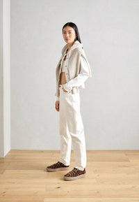 Cardigan court beige sur un top court blanc, accompagné d'un jean taille haute blanc et de baskets marron à imprimé léopard. Matériaux texturés visibles.