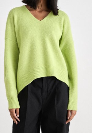 Femme portant un pull en maille vert citron à col en V avec manches longues, assorti à un pantalon noir ample sur un fond uni.