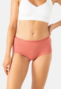 Femme portant un soutien-gorge de sport blanc et un sous-vêtement taille haute corail, se tenant avec une main sur la hanche devant un fond uni.