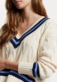 Krämfärgad kabelstickad tröja med V-hals, som har marinblå och ljusblå randiga detaljer på kragen och ärmarna.