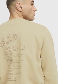 Jasno beżowy sweatshirt z okrągłym dekoltem. Tył zdobi nadrukowany wzór „Bucket List” z miejscami podróży i aktywności w kolorze brązowym.
