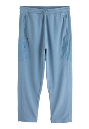 Nahaufnahme einer Person, die hellblaue Jogginghosen mit Tunnelzugbund und seitlicher Reißverschlusstasche trägt, stehend mit entspannter linker Hand.