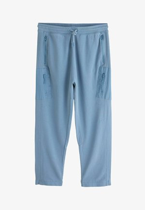 Prim-plan cu o persoană purtând pantaloni de trening bleu cu șnur în talie și buzunar lateral cu fermoar, stând cu mâna stângă relaxată.