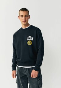 Sort sweater med ribbede manchetter og kant. Har hvid tekst "THE STONE ROSES" og en gul grafisk illustration på brystet. Lavet af bomuldsstof.