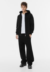 Črna zip-up kapuca s sprednjimi žepi, nošena preko bele majice, v kombinaciji s črnimi trenirkami in črnimi športnimi čevlji.