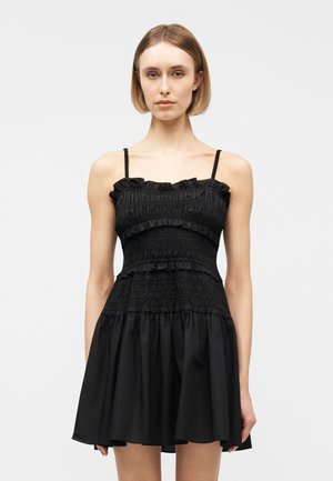 Kobieta o krótkich blond włosach, ubrana w czarną mini sukienkę na cienkich ramiączkach z marszczonym przodem, stojąca na tle jasnej, jednolitej ściany.