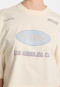 Krótki rękaw koszulki w kolorze kremowym z napisem "GUESS Originals" w kolorze niebieskim oraz "LOS ANGELES, CA" w kolorze czerwonym. Wykonana z miękkiego materiału bawełnianego.