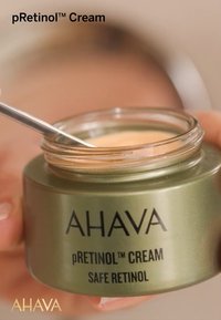 Tarro verde de crema pRetinol™ con tapa transparente, etiquetado con "AHAVA" y detalles del producto. Una espátula plateada está suspendida sobre la crema.