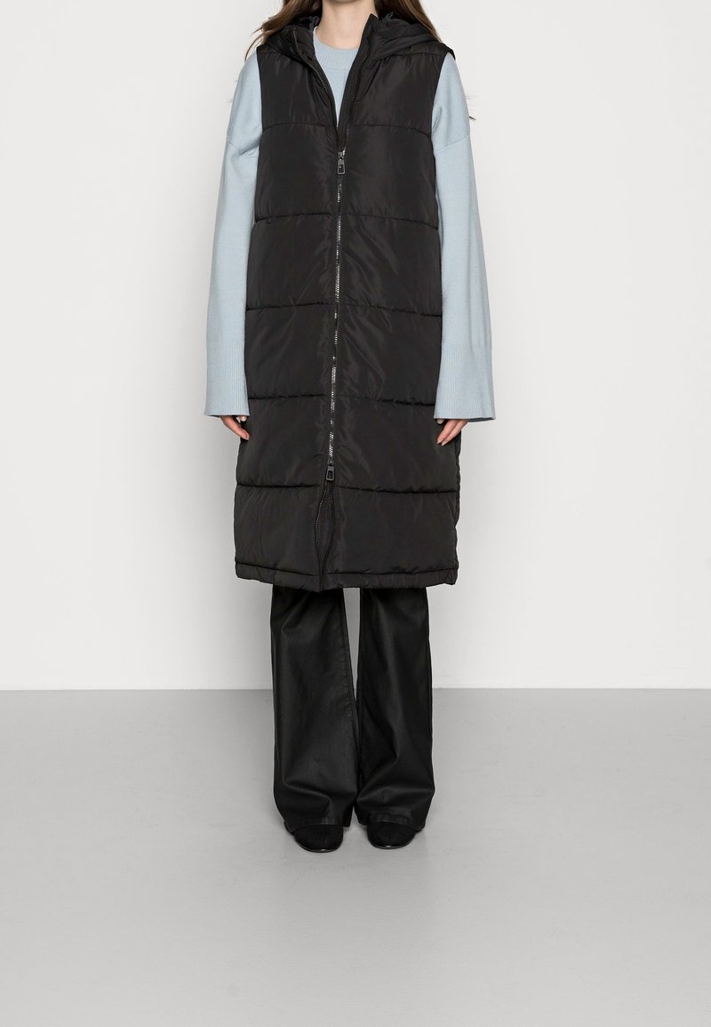 Czarna pikowana kamizelka z wysokim kołnierzem, zamkiem z przodu i pikowanym wzorem. Noszona na jasnoniebieską bluzkę z długim rękawem i czarne, szerokie spodnie.