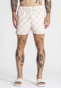 Mies, joka pukeutunut beigen ja valkoisen ruudullisiin shortseihin ja niihin sopiviin liukujiin, seisoo valkoista taustaa vasten. Hänen käsivarsissaan ja jaloissaan näkyy tatuointeja.