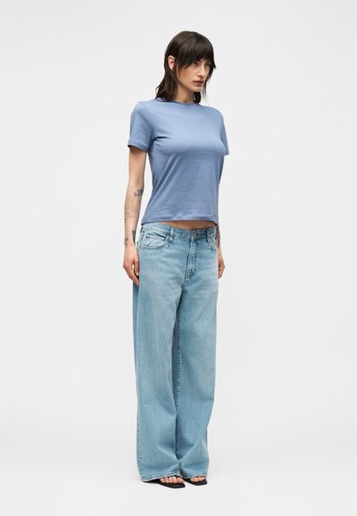 Γυναίκα που στέκεται φορώντας ένα ανοιχτό μπλε κοντομάνικο μπλουζάκι, ανοιχτό μπλε τζιν παντελόνι με φαρδιά μπατζάκια και μαύρα σανδάλια με ανοιχτά δάκτυλα σε άσπρο απλό φόντο.