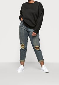 Osoba w czarnej długiej bluzie z długim rękawem, w zniszczonych, podwiniętych niebieskich dżinsach z przetarciami na kolanach i białych trampkach, stojąca z jedną ręką w kieszeni.