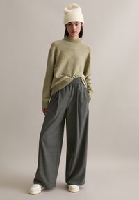 Pull tricot vert olive avec col montant et coupe décontractée, assorti à un pantalon large à carreaux gris et des baskets blanches. Bonnet crème.